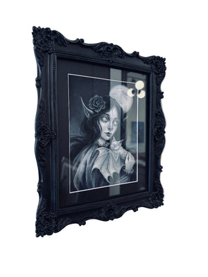 A framed black and white gothic art print featuring a woman with a rose and bat wings, enclosed in an ornate black frame and mounted on a white wall.