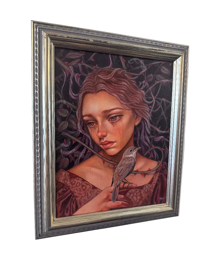 A framed fantasy portrait painting of a woman with reddish-brown hair and a small bird on her shoulder, set in an ornate silver frame and hanging on a white wall.
