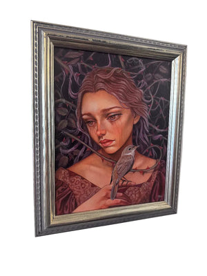 A framed fantasy portrait painting of a woman with reddish-brown hair and a small bird on her shoulder, set in an ornate silver frame and hanging on a white wall.
