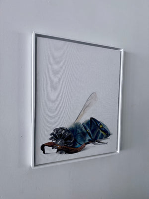 A framed art print featuring a detailed illustration of a blue and black bee on a white background, hung on a light gray wall.