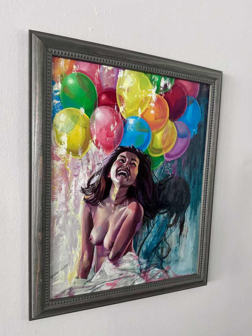 A framed painting on a white wall, depicting a laughing woman with dark hair surrounded by colorful balloons in green, yellow, pink, blue, orange, and purple, enclosed in a dark gray frame.