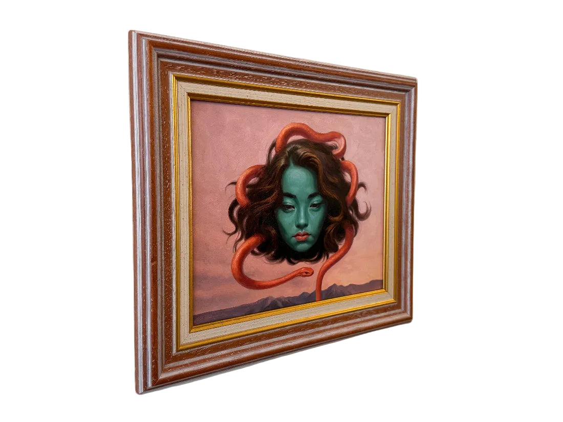 A framed surreal painting hanging on a beige wall, featuring a green-skinned woman with brown hair wrapped in an orange-red snake against a pink sky background.