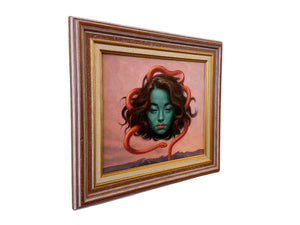 A framed surreal painting hanging on a beige wall, featuring a green-skinned woman with brown hair wrapped in an orange-red snake against a pink sky background.