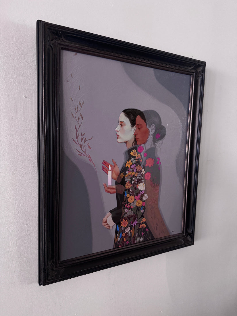A framed painting featuring a stylized portrait of two women in profile, one in a floral patterned dress, hanging on a plain white wall.