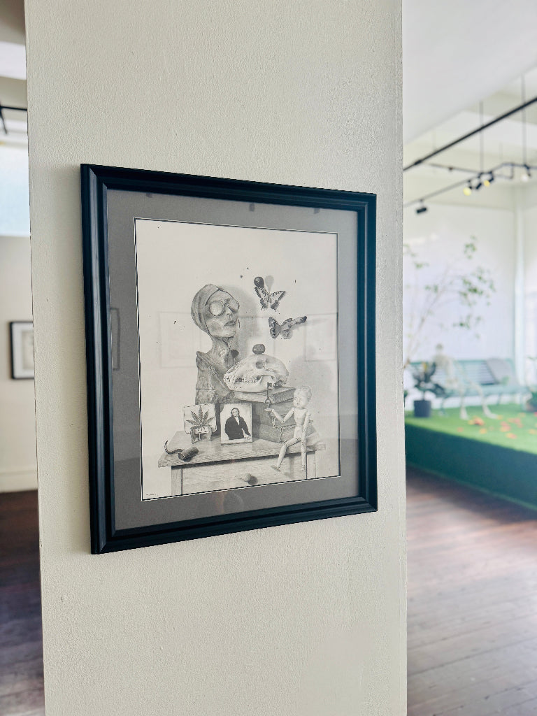 Framed black and white artwork on a wall with a view of a gallery. 