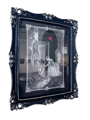 A framed black and white ink drawing featuring a Victorian-era woman in a gown near an ornate gothic archway, with a small pink bird in the sky. The print is housed in an ornate black frame with silver beading and is mounted on a white wall.
