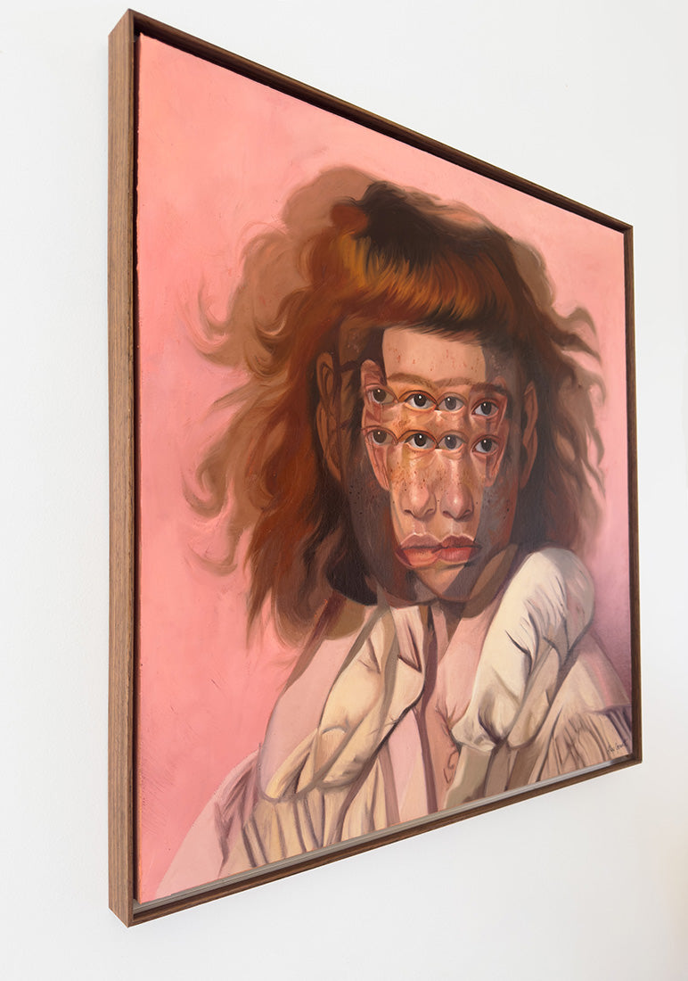 A framed portrait painting of a woman with multiple eyes on her face, set against a pink background, hanging on a white wall.