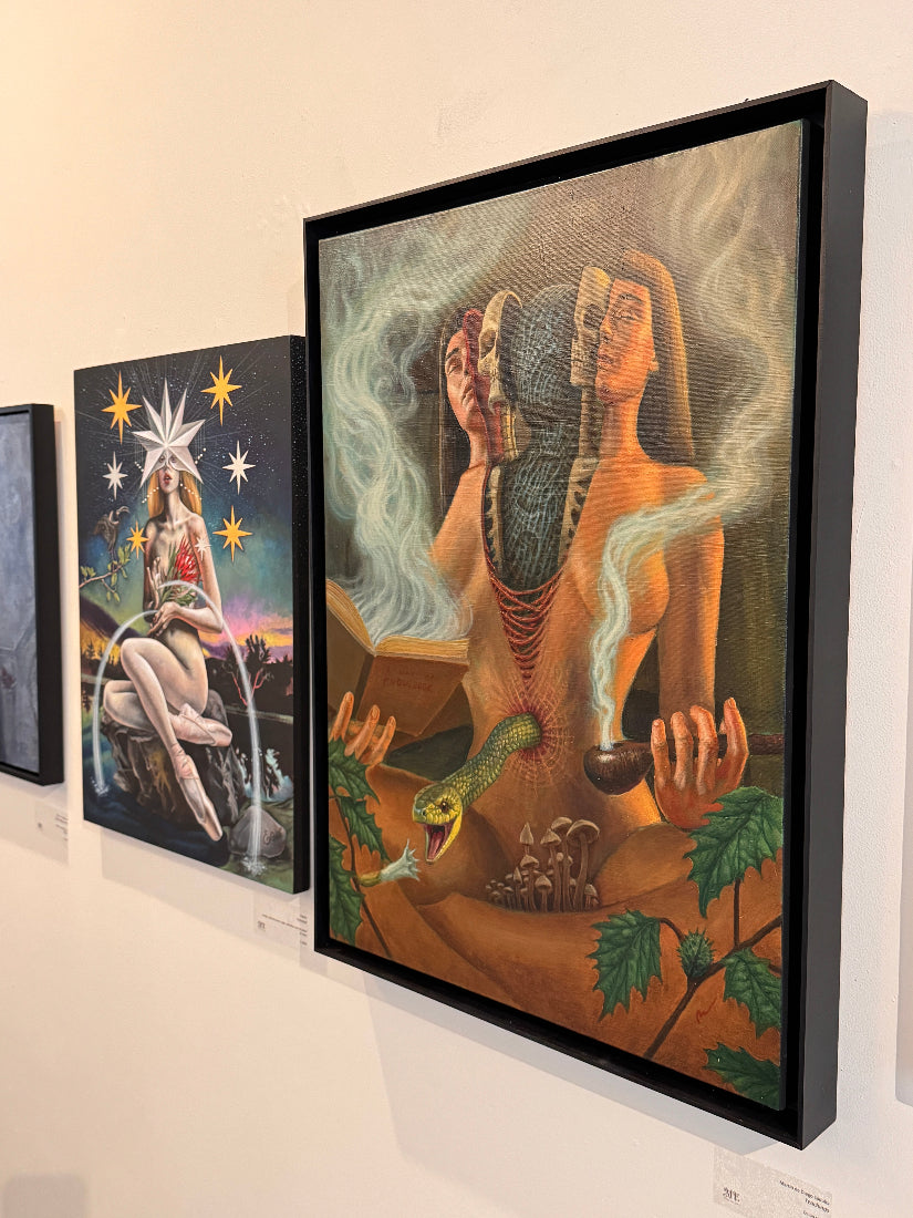 A large framed surreal painting hangs on a gallery wall. The artwork depicts a seated nude female figure with a snake wrapped around her torso, smoke-like forms, stars, and a small snake head in the foreground.