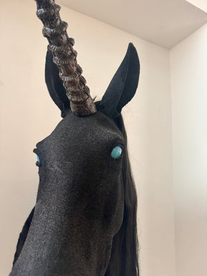 Unicorn head sculpture with dark gray body and black mane against a plain wall.