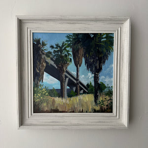 Framed painting of palm trees and a bridge on a white wall