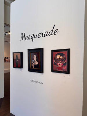 An art gallery wall with the word 'Masquerade' in black script above three framed portrait artworks. The center framed piece is labeled 'The Portrait Show 14.