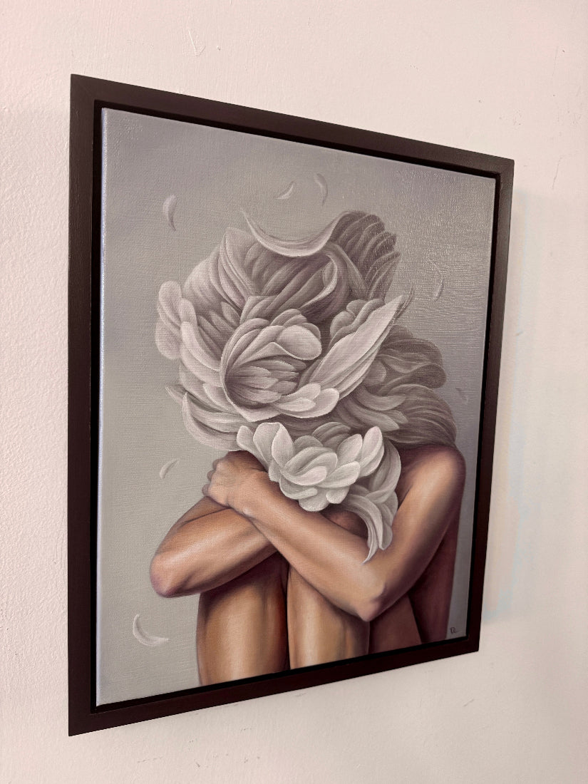 A framed painting hanging on a white wall, showing a stylized figure with arms crossed and large white flowers forming the head, rendered in soft gray, beige, and brown tones with a dark frame.