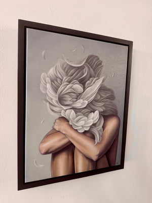 A framed painting hanging on a white wall, showing a stylized figure with arms crossed and large white flowers forming the head, rendered in soft gray, beige, and brown tones with a dark frame.