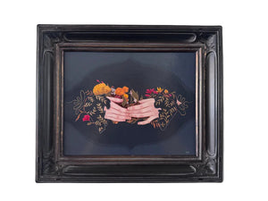 A framed painting featuring a dark illustration of two hands holding a bouquet of orange and red flowers, set within an ornate black frame and mounted on a light gray wall.