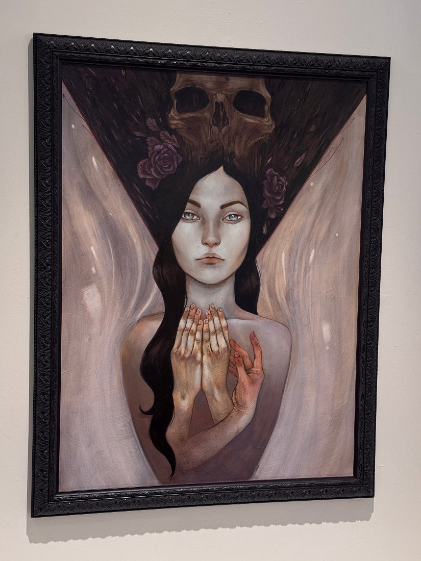 A framed surreal painting hanging on a light wall. The artwork shows a pale woman with long dark hair, clasped hands, a large dark triangular headdress with a skull and roses, and soft pinkish-white wings or drapery behind her.