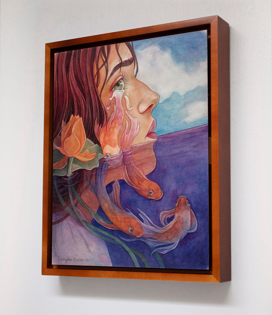 A framed watercolor painting on canvas, shown hanging on a light gray wall. The artwork features a woman's profile with flowing reddish-brown hair, an orange flower, orange fish, and a blue-purple landscape under a cloudy sky.
