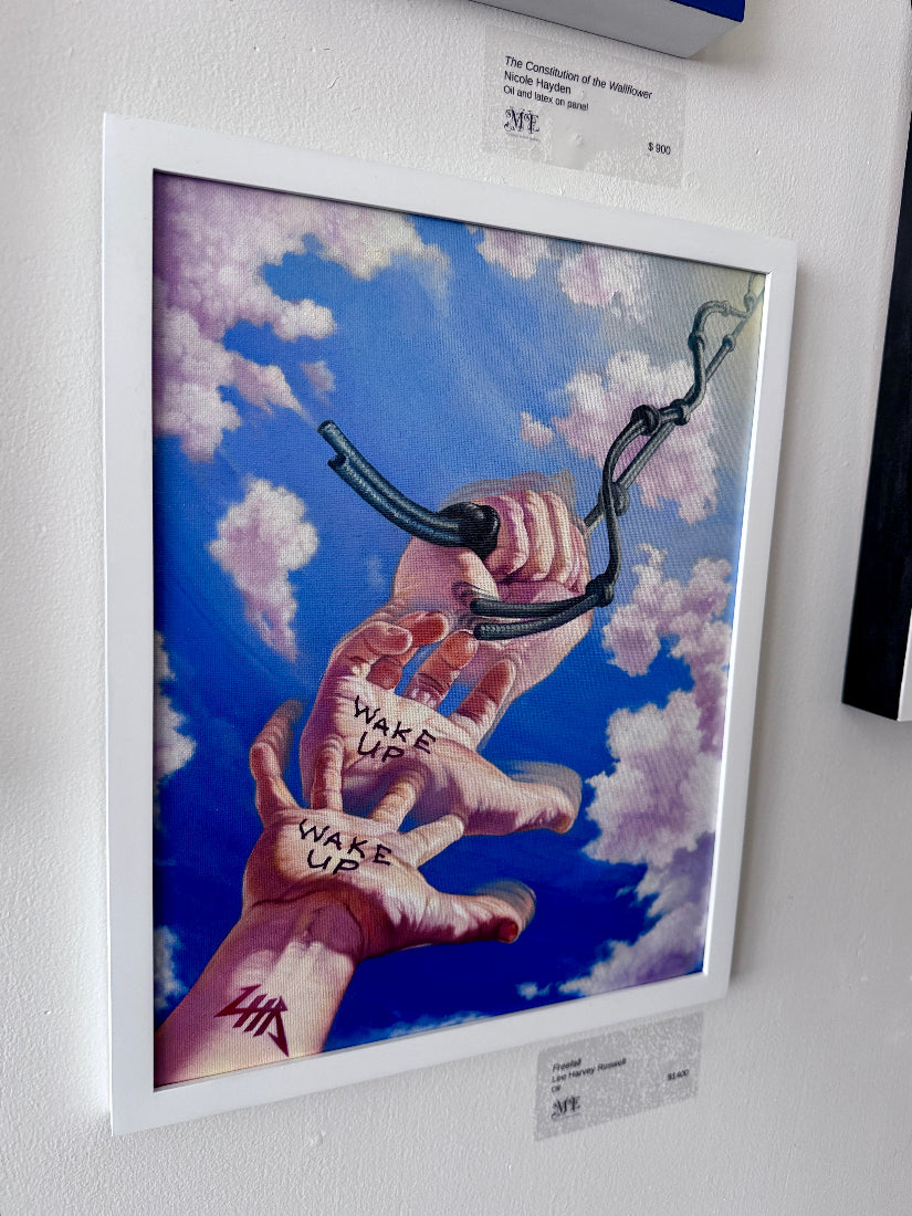 A framed painting hanging on a white wall, depicting two hands reaching up through blue sky and clouds, with one hand holding a black chain and the words 'WAKE UP' written on the other hand.