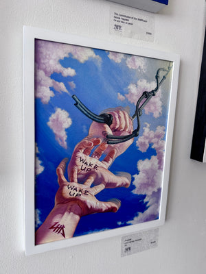 A framed painting hanging on a white wall, depicting two hands reaching up through blue sky and clouds, with one hand holding a black chain and the words 'WAKE UP' written on the other hand.