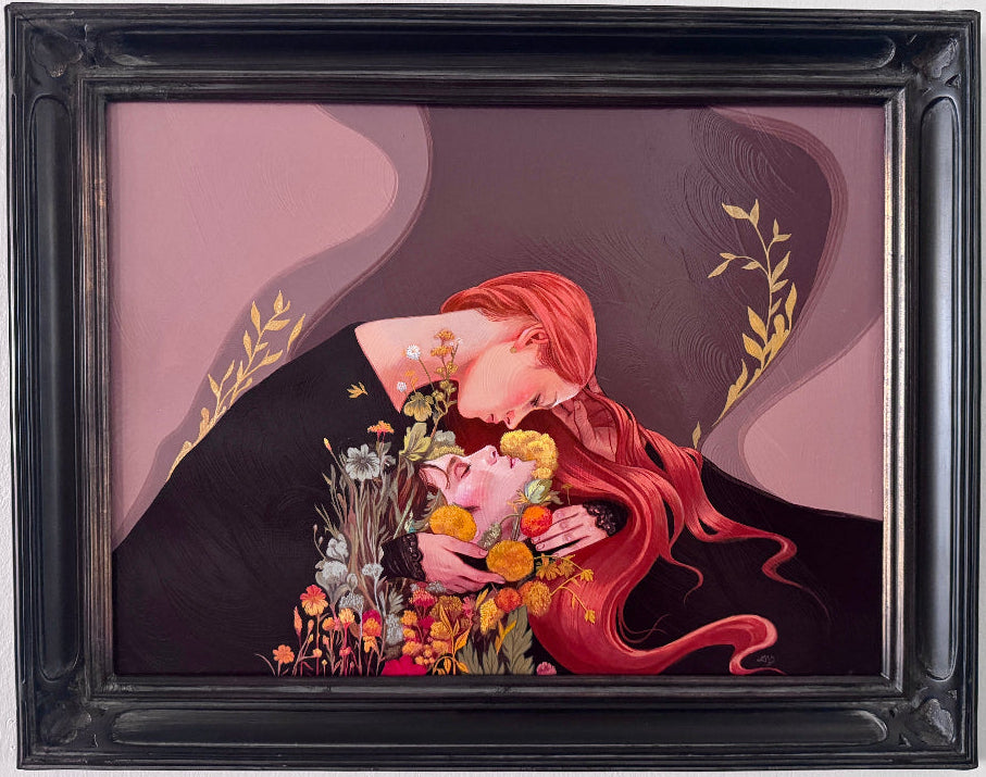 A framed painting featuring a stylized illustration of a woman with long red hair kissing a person, with flowers and gold leaf details, housed in a black ornate frame against a white wall.