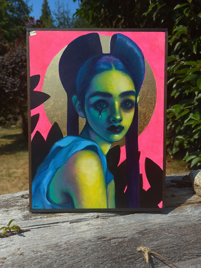 A framed painting featuring a stylized portrait of a woman with green skin, blue hair, and a bright pink background, displayed outdoors on a weathered wooden surface with trees in the background.