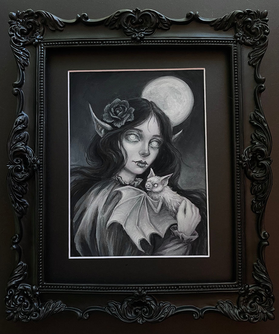 A framed gothic art print featuring a black and white illustration of a vampire woman holding a bat, with a rose in her hair and a full moon in the background, set in an ornate black frame.