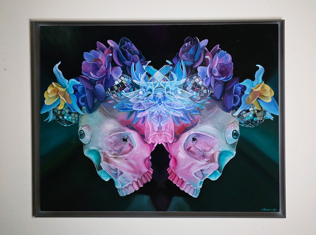 A framed surreal painting featuring two mirrored pink and blue skulls adorned with purple flowers, glowing blue crystals, and small disco-ball-like accents against a dark background, displayed on a light wall.