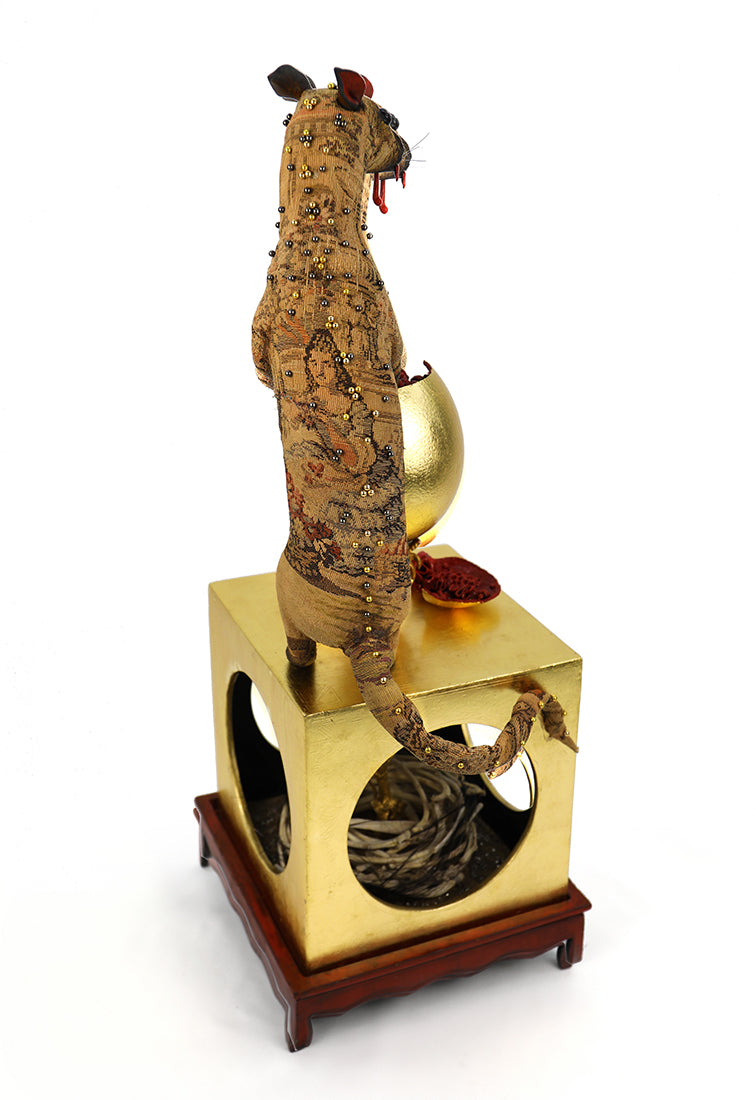 A surreal figurative art sculpture featuring a standing deer holding a goblet of red liquid, positioned on top of a gold pedestal with a circular cutout showing gray twigs, set against a plain white background.