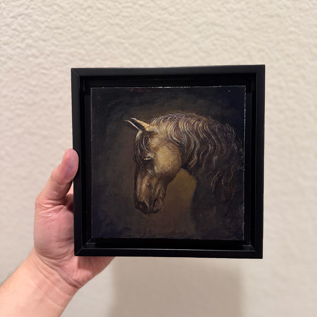 Framed painting of a horse held by a hand against a plain background