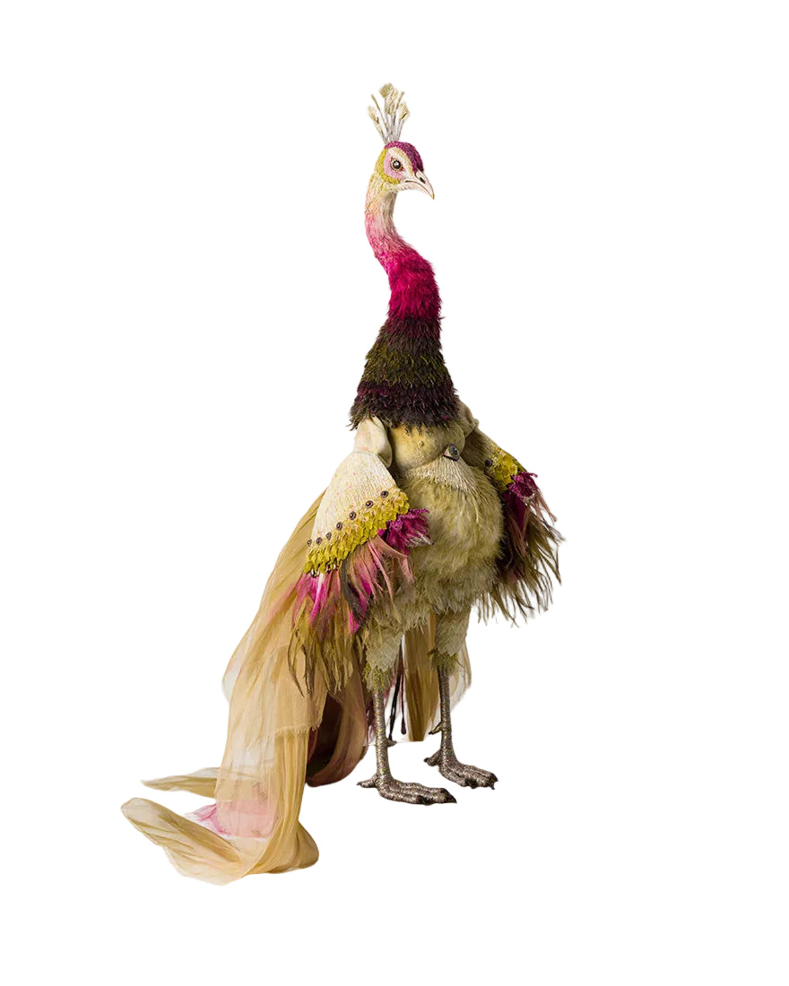 A whimsical, handcrafted peacock sculpture or costume stand featuring a long flowing beige feathered tail, a dark brown textured neck, a vibrant magenta-pink head and chest, and yellow accents on the wings, mounted on a round wooden base with small wheels against a plain light gray background.