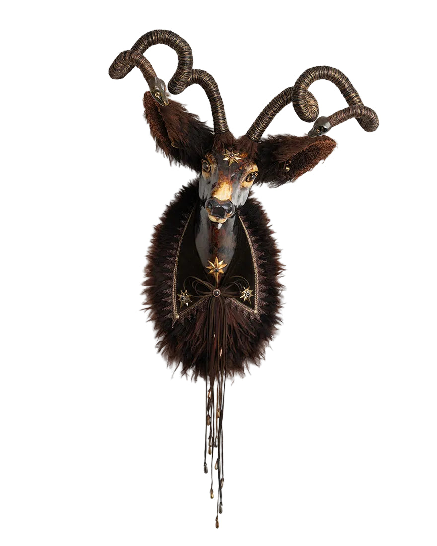 A decorative taxidermy-style wall mount featuring a dark brown goat or ram head with curled horns, dark fur, and gold-toned embellishments on a plain white background.