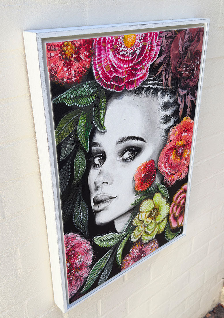 Framed artwork of a woman's face surrounded by colorful flowers on a white wall.