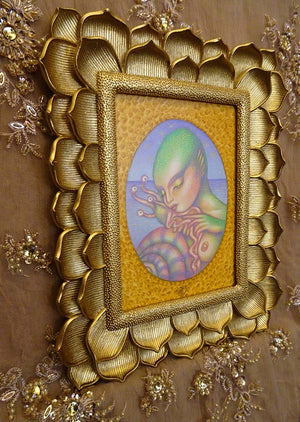 A framed art print featuring a stylized green-skinned alien on a textured gold background, enclosed in an ornate gold lotus-shaped frame and mounted on a brown embroidered fabric backdrop.